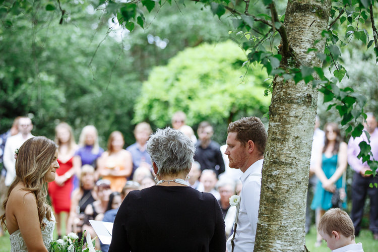 Behind the celebrant, Mitch in braces smiles at Elysha beside a birch trunk.