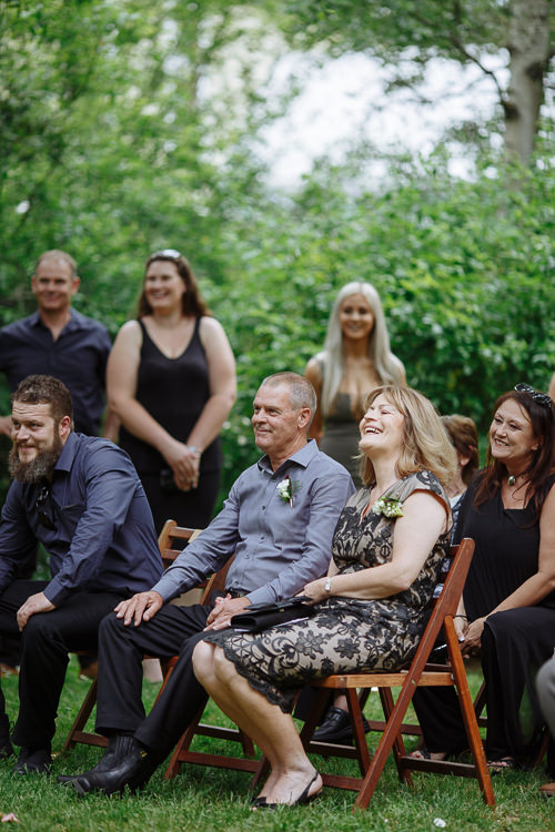 Seated guests laugh on timber folding chairs, a woman in black lace beside a man with a boutonniere.