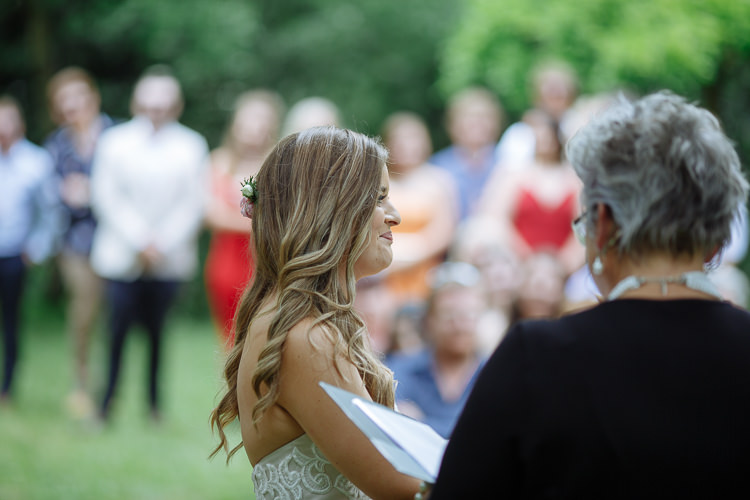 Elysha in profile smiles at the celebrant holding a folder, guests blurred behind.