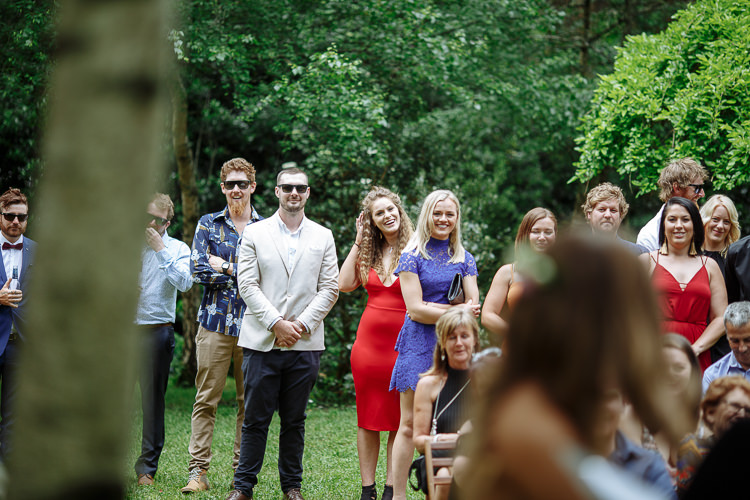 Standing guests watch the ceremony, a woman in red lace with a hand to her cheek among them.