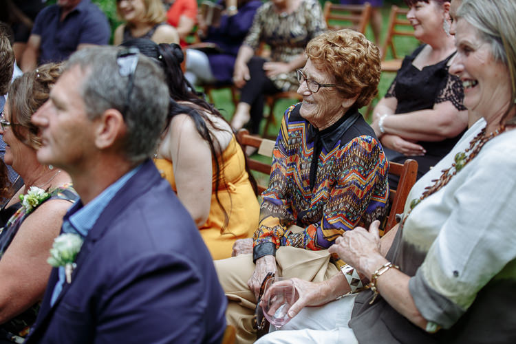 An older woman in a bright patterned jacket laughs among the seated guests during the ceremony.