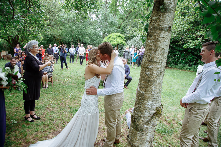 The first kiss beside a birch trunk as the celebrant claps and guests stand across the lawn.