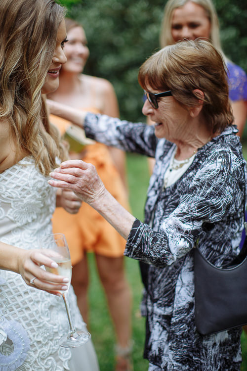 An older woman in sunglasses and a patterned jacket reaches to hug Elysha holding a champagne flute.