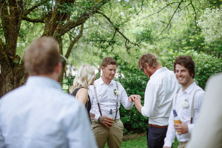 Under a tree, Mitch shakes hands with a guest while a groomsman laughs with a beer.
