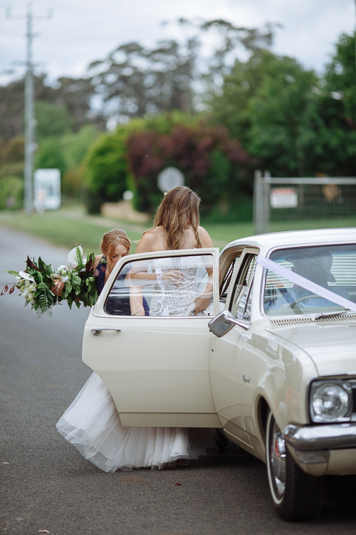 On a country road, Elysha climbs into the ribboned cream sedan as a bridesmaid helps her.