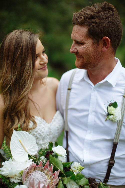 Close on the couple smiling at each other, the anthurium and king protea bouquet between them.