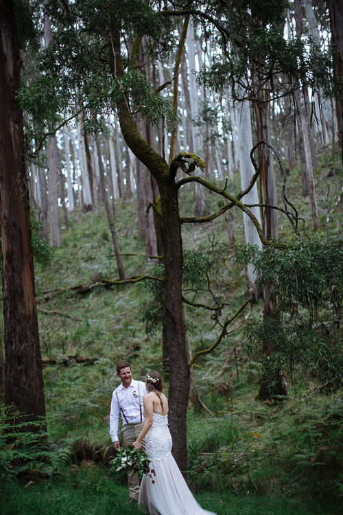 Beneath a mossy branching tree, Mitch smiles at Elysha holding her bouquet in the forest.