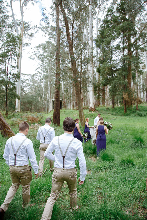 From behind, three groomsmen in braces walk through long grass toward the bridesmaids and the couple.