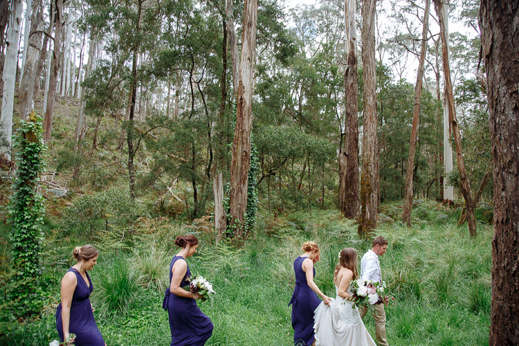 The bridal party walks in a line through long grass beneath tall gums, Mitch last.
