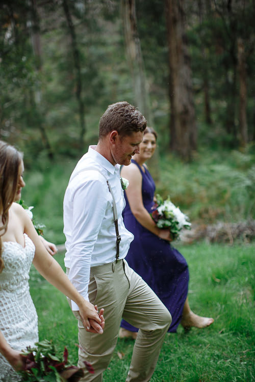 Mitch leads Elysha by the hand through the grass, a barefoot bridesmaid in navy behind.