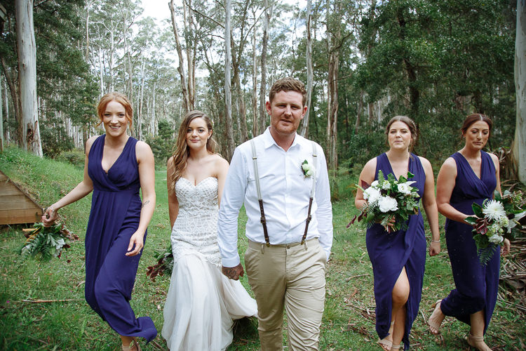 Elysha and Mitch walk toward camera hand in hand with three bridesmaids in navy, gum forest behind.