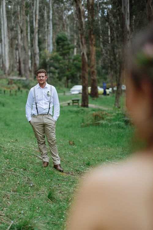 Over Elysha's blurred shoulder, Mitch waits in the clearing with hands in his pockets.