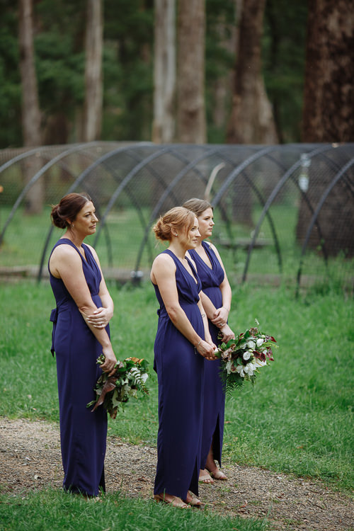 Three bridesmaids in navy with foliage bouquets stand before a row of wire hoops beneath tall gums.