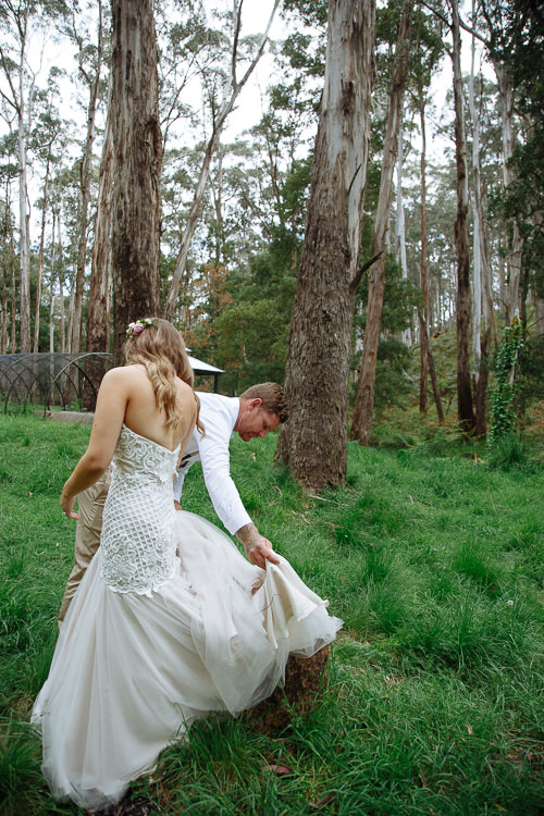 Mitch lifts Elysha's tulle train from the grass as she walks ahead, bare back to camera.