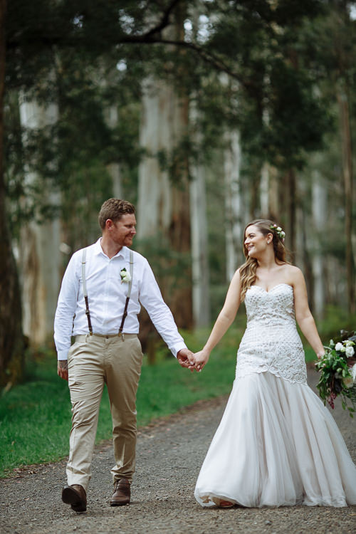 Elysha and Mitch walk hand in hand down a gravel forest road smiling at each other.