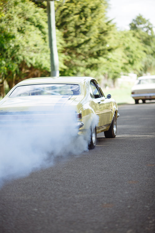 The yellow vintage coupe lays a cloud of tyre smoke as it pulls away, the cream sedan behind.