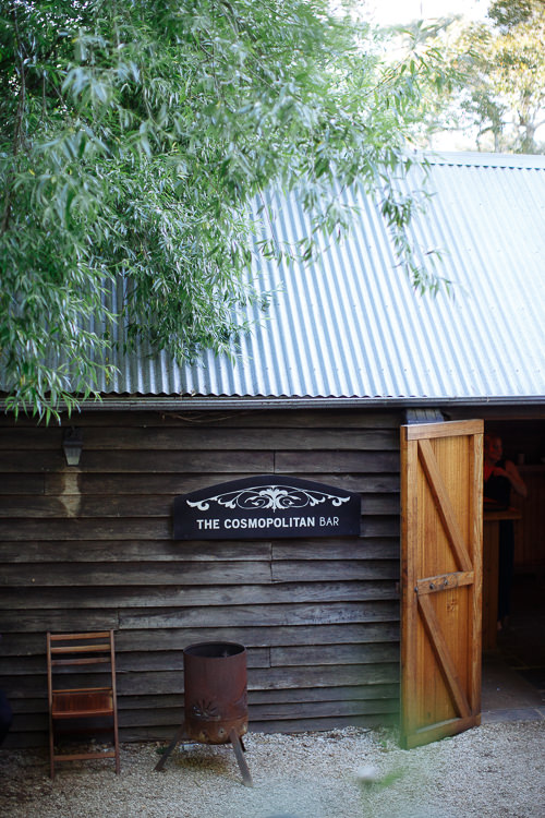 A sign lettered The Cosmopolitan Bar on a timber outbuilding beside a rusted brazier and open barn door.