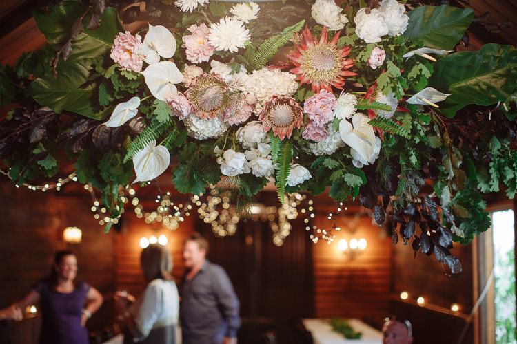 A hanging installation of king protea, anthurium and foliage above fairy lights in the timber room.