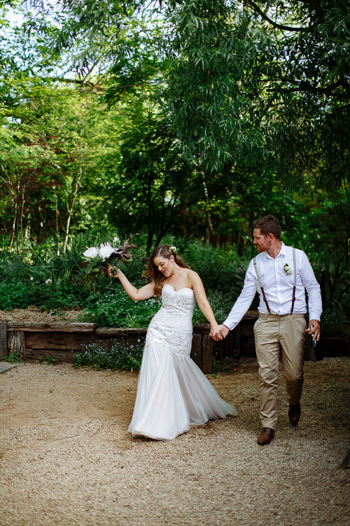 Elysha swings her bouquet high, laughing, as Mitch leads her by the hand under a willow.