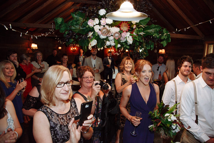 Guests cheer beneath the hanging florals in the timber room at The Cosmopolitan Hotel, phones raised.