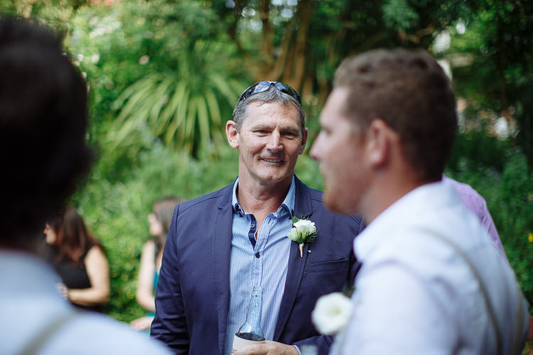 A man in a navy jacket with a white boutonniere smiles in the garden.