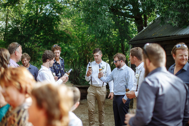 Groomsmen and guests with beers gather on the gravel beside the black timber Cosmopolitan bar under willows.
