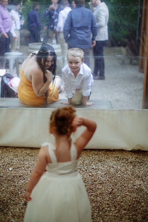 Through glass, a flower girl watches a boy and a woman in yellow kneeling on a low stage.