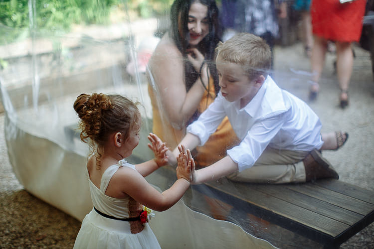 The flower girl and a boy press their palms together through the glass, laughing.