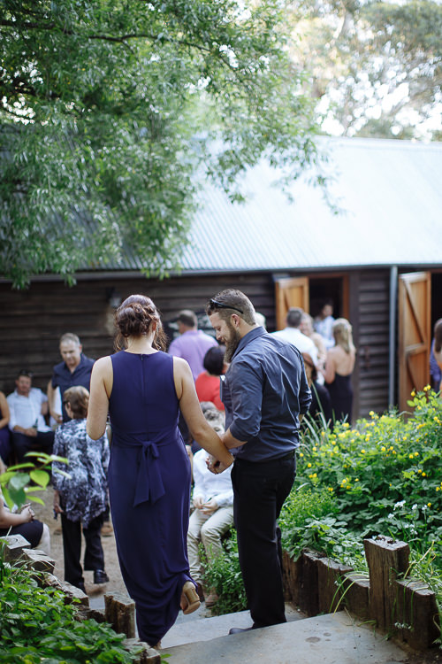 From behind, a bridesmaid in navy and a bearded man walk hand in hand toward the barn doors.