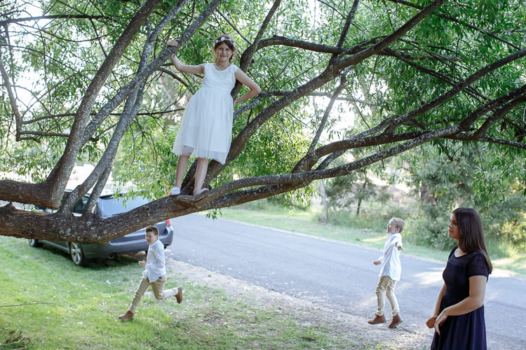A girl in a white dress stands on a low willow branch while two boys run below.