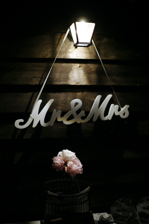 A white Mr and Mrs sign hangs by ribbon beneath a lantern above pink peonies in a birdcage.