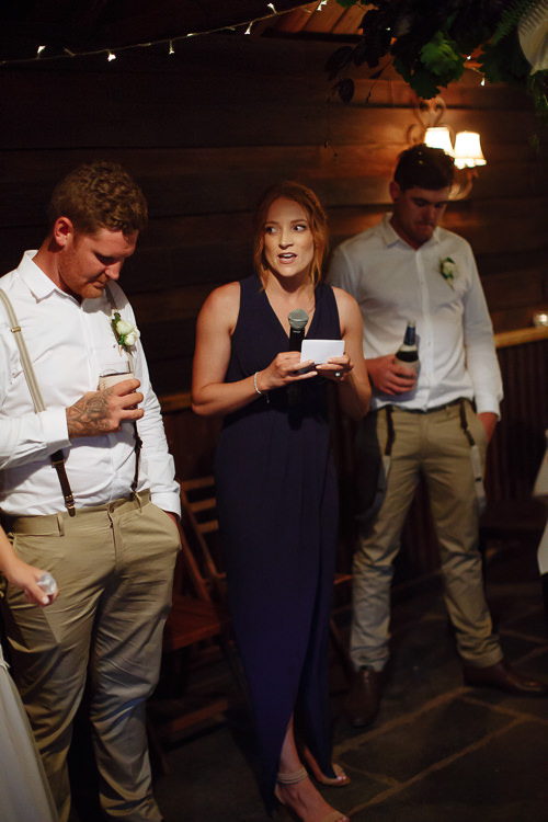 A bridesmaid in navy reads her speech from a card between Mitch in braces and a groomsman.