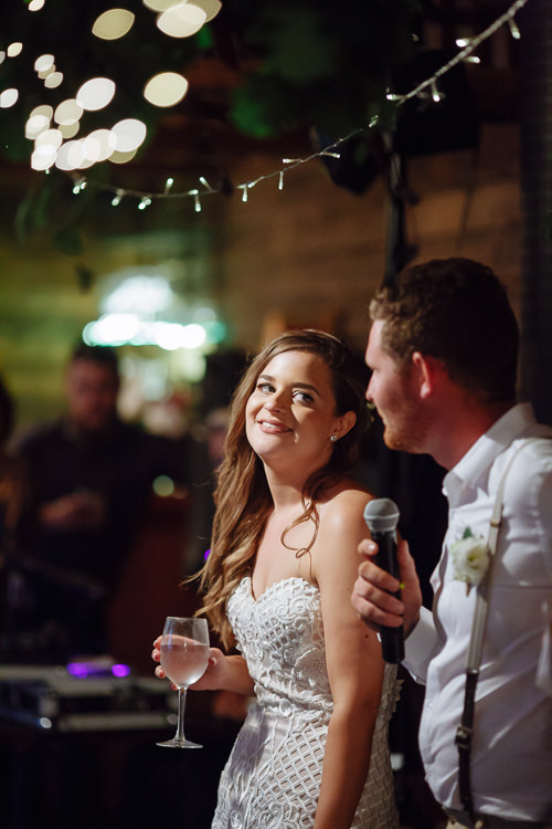 Elysha smiles back at Mitch over her shoulder as he speaks, fairy lights above.