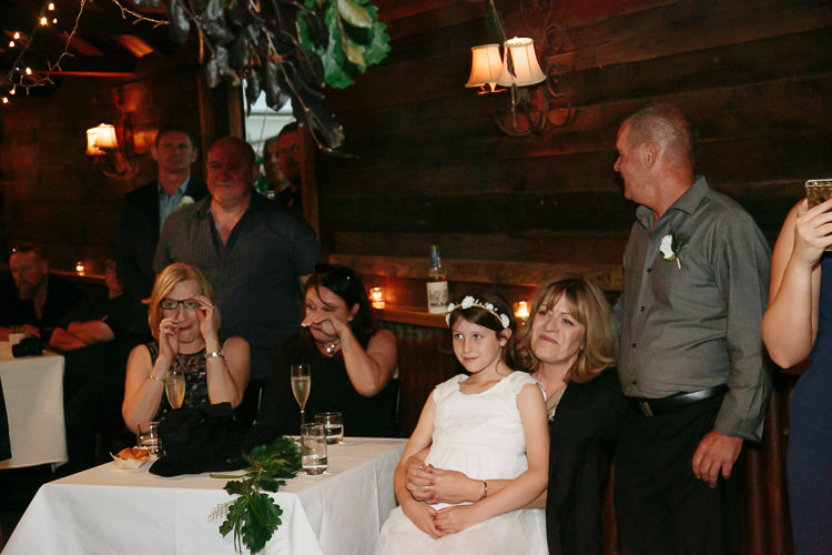 Guests wipe tears during the speeches, a flower girl in a flower crown on a woman's lap.