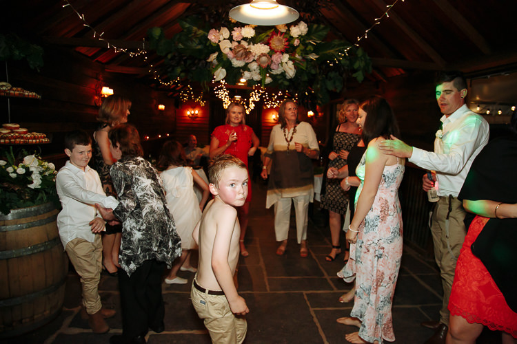 A shirtless boy dances at the front of the stone floor as guests dance beneath the hanging florals.