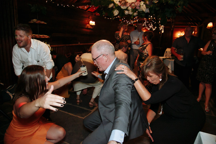 A grey suited man crouches mid dance between a laughing woman in orange and a woman in black.