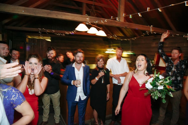 Guests cheer under festoon lights, a woman in red holding the caught bouquet at right.