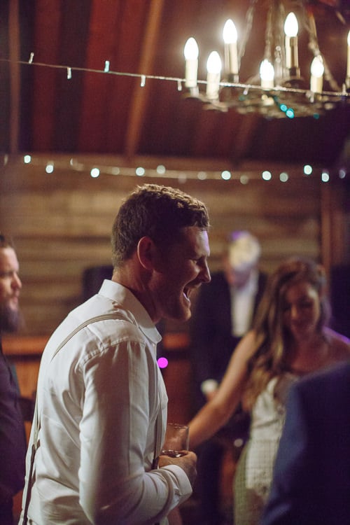 Laughing in profile beneath a candle chandelier and fairy lights, Mitch holds a glass.