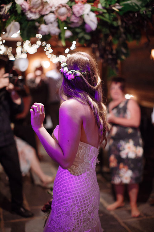 From behind, Elysha dances lit purple beneath the hanging florals, a guest filming at left.