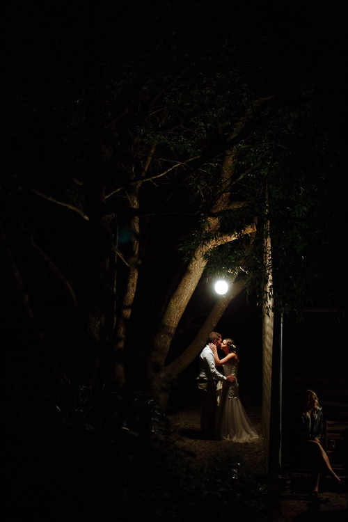 The couple kiss beneath the lone globe among dark branches at night, the tulle skirt catching light.
