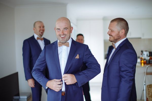 Groom laughing in his navy suit and wooden bow tie with his groomsmen