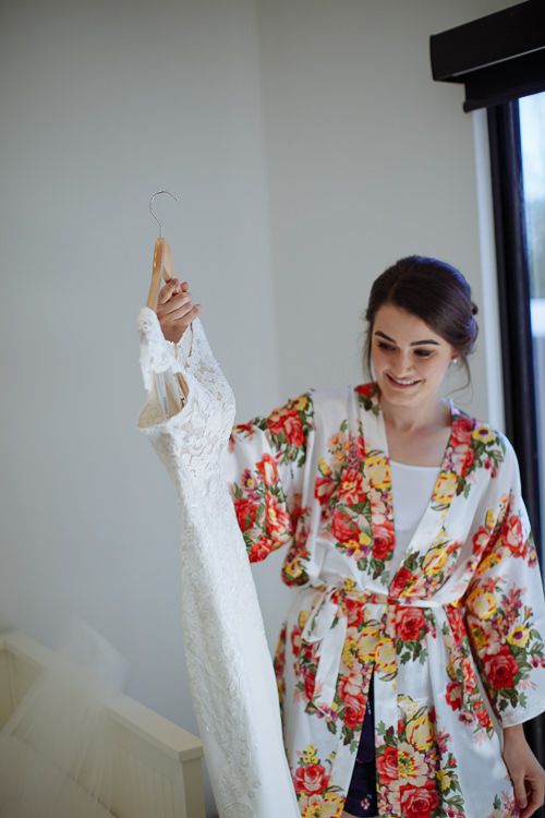 Bride in her floral robe holding the lace gown on its hanger