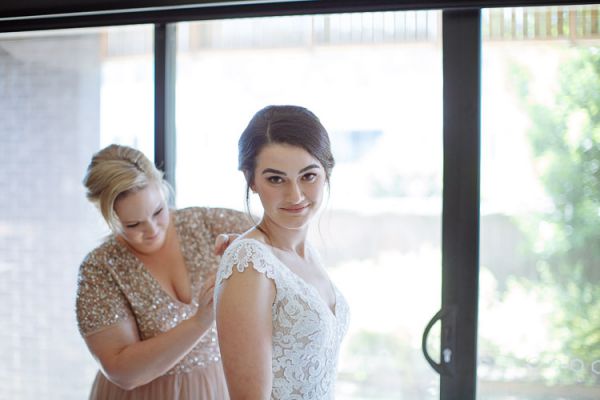 Bridesmaid helping the bride into her gown by the window