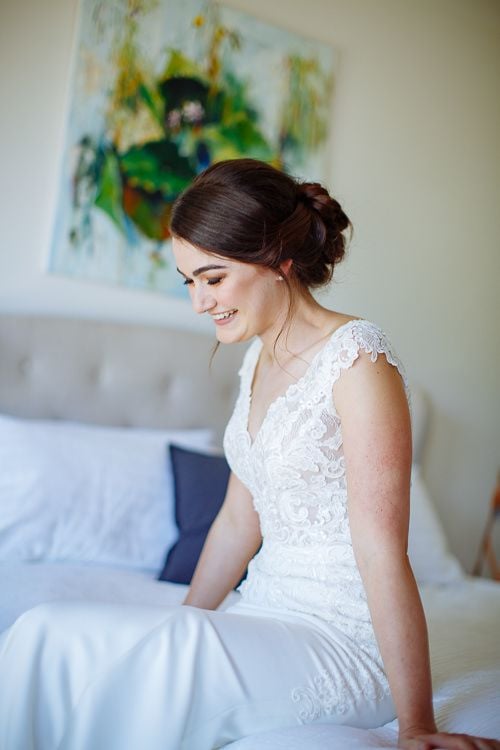 Bride smiling softly in her lace gown beside the bed
