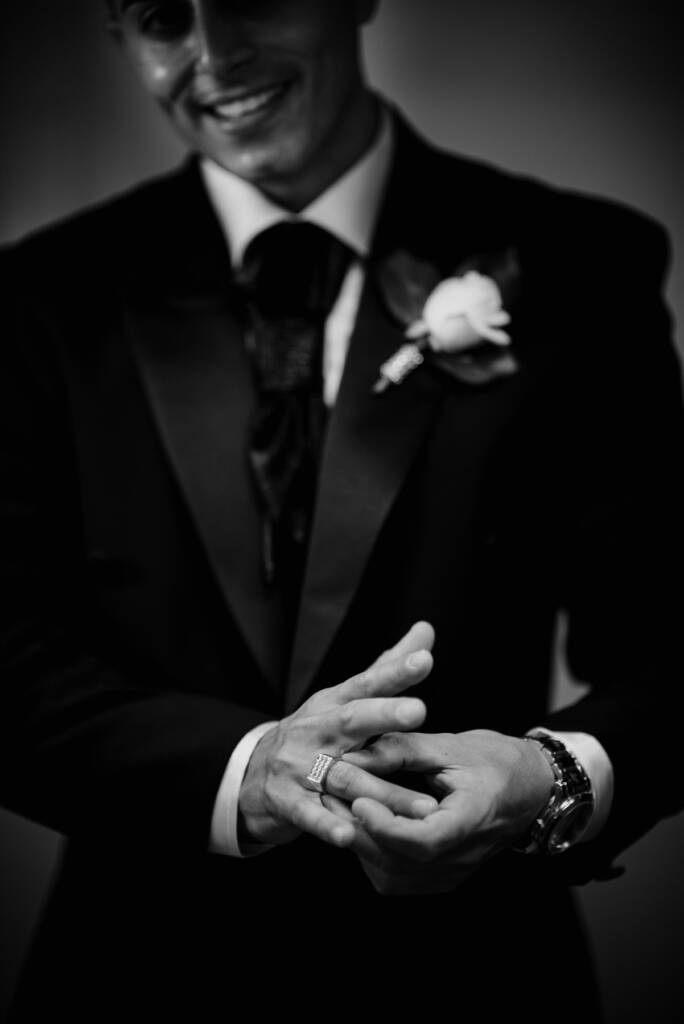 Michael adjusts his ring beside his cravat and boutonniere in a black-and-white detail photo