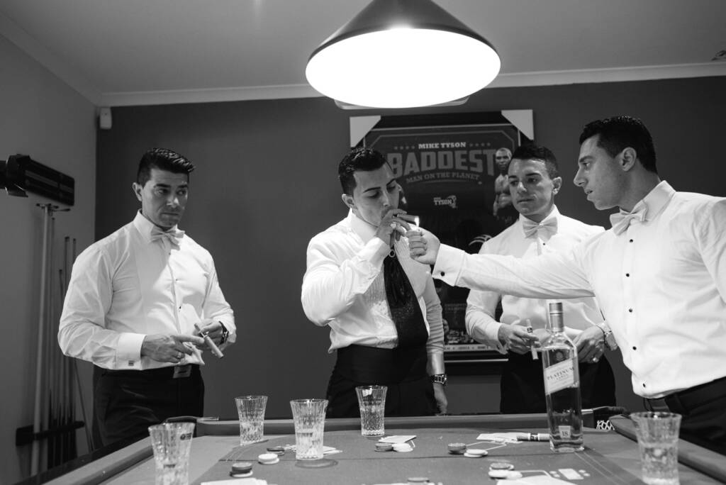 A groomsman lights Michael's cigar while friends gather around a card table