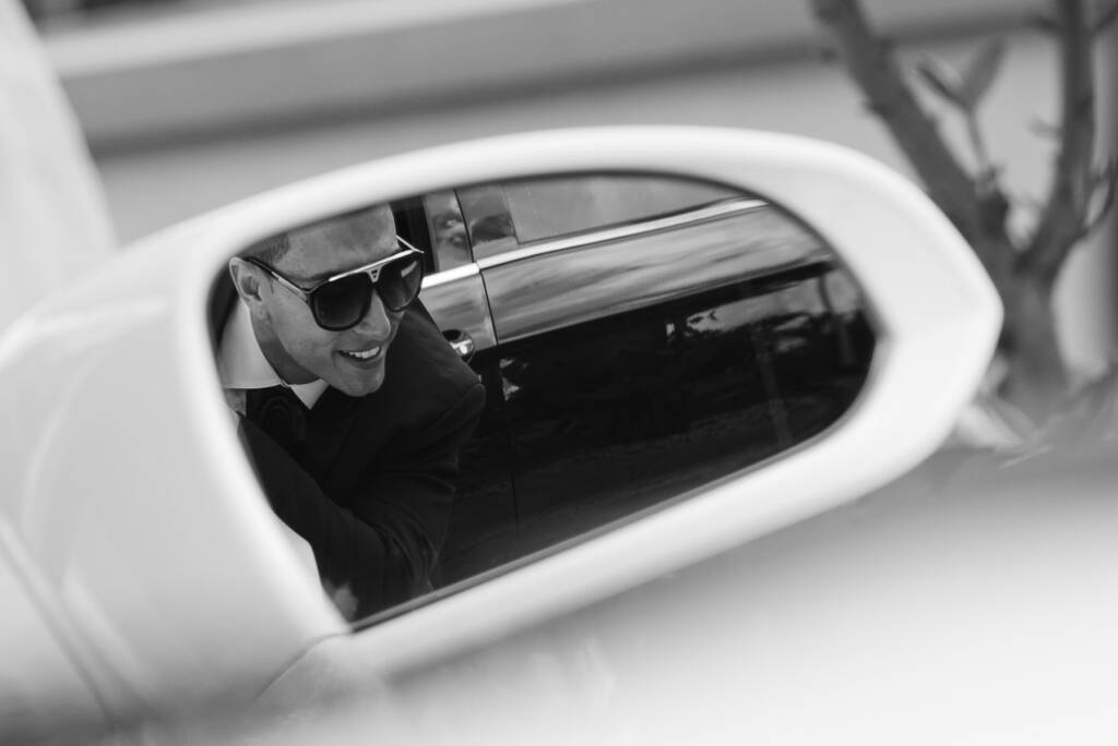 A groomsman smiles in the wing mirror of a white convertible
