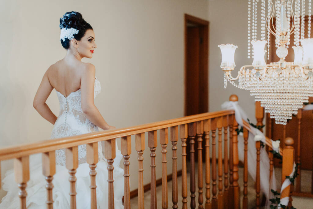 Danielle stands on the staircase landing in her lace gown beside a chandelier