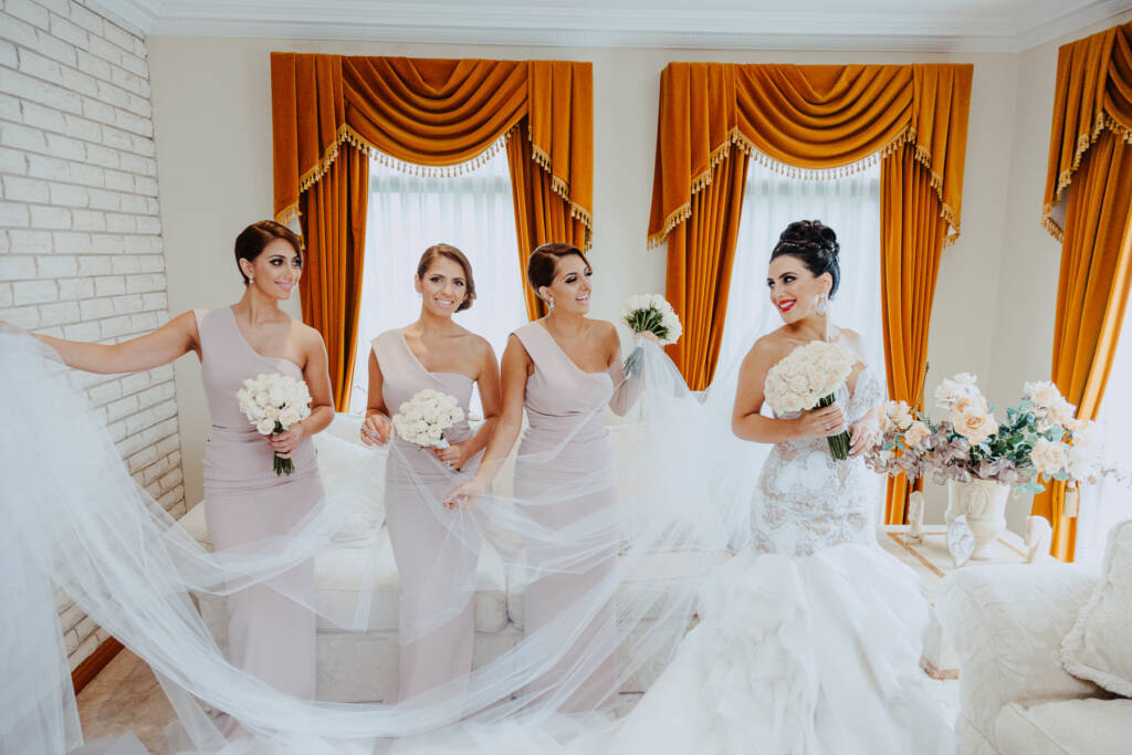 Three bridesmaids hold Danielle's veil out around her during bridal preparation