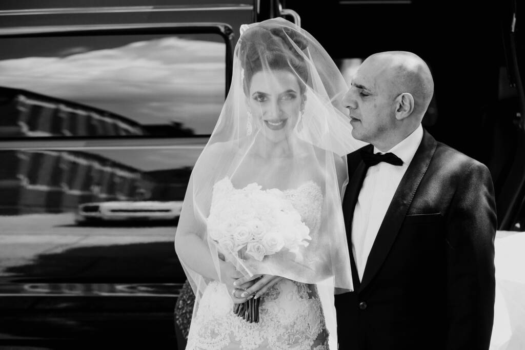 Danielle smiles beneath her veil beside a suited man in a black-and-white arrival photo